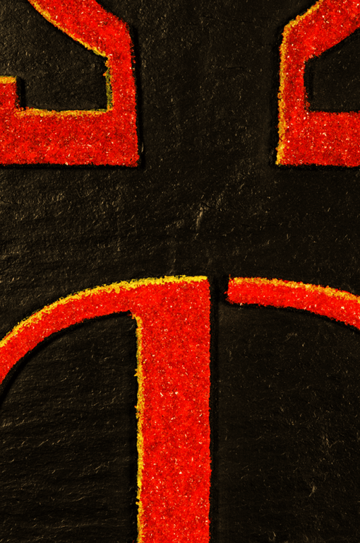 Extrem detaillierte Nahaufnahme des unteren Teils des Schiefer-Reliefbildes "Alpha und Omega", die die feinen Strukturen des roten Kunstsandsteins und die Schnittpunkte der stilisierten Formen und des angedeuteten Kreuzes zeigt.