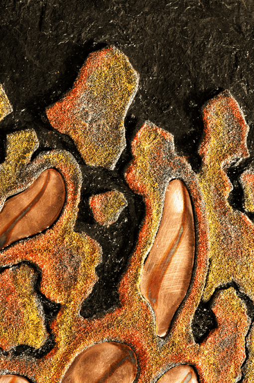Nahaufnahme eines Abschnitts des Schiefer-Reliefbildes "Eruption", die die feinen Texturen des dunklen Schiefers, die körnigen Oberflächen des Kunstsandsteins in glühenden Farben und die geschliffenen Details der Kupferelemente zeigt.